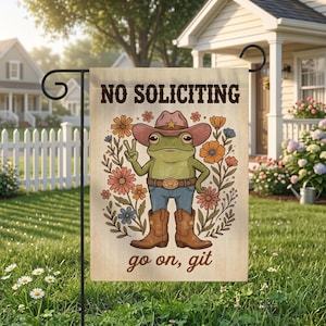 May include: A garden flag with a frog wearing a cowboy hat, boots, and jeans, surrounded by flowers. The flag reads "NO SOLICITING" and "go on, git". The frog is giving a peace sign.