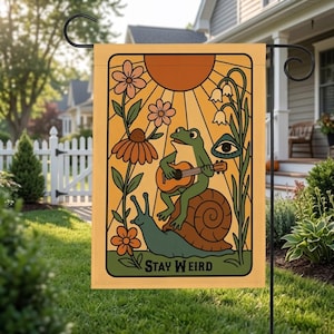 Puede incluir: Una bandera de jardín con un diseño caprichoso. Una rana verde toca la guitarra mientras monta un caracol, rodeada de flores y un sol. El texto "STAY WEIRD" está en la parte inferior, todo sobre un fondo amarillo.