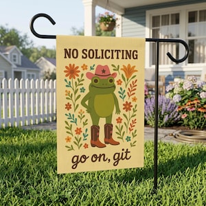 May include: A garden flag with a frog wearing a cowboy hat and boots, surrounded by flowers. The flag reads "NO SOLICITING" above the frog and "go on, git" below. The flag is displayed on a black metal stand in a yard.