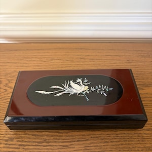 May include: A rectangular, lacquered box with a dark red frame and black base. The lid features an oval inlay with a white bird and floral design. The bird has a long tail and is surrounded by delicate, colorful accents. The box is set on a wooden surface.