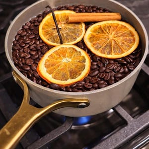 May include: A saucepan on a hob filled with coffee beans, orange slices, a cinnamon stick, and a vanilla pod. Steam rises from the pan, indicating it is heated. The pan has a gold handle.