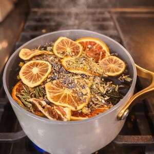 May include: A light gray pot with gold handles on a stovetop, filled with simmering water and dried orange slices, herbs, and lavender. Steam rises from the pot, suggesting a fragrant infusion or potpourri.