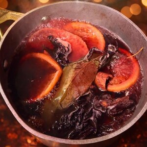 Könnte beinhalten: Ein Edelstahltopf mit köchelndem Glühwein. Orangenscheiben, Lorbeerblätter und andere Gewürze schwimmen in der dunkelroten Flüssigkeit. Dampf steigt aus dem Topf auf, und der Hintergrund ist verschwommen mit warmen, goldenen Bokeh-Lichtern.