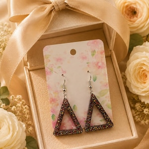 Puede incluir: Pendientes colgantes triangulares con un diseño brillante y multicolor. Se exhiben en una caja de regalo dorada abierta con un lazo a juego. El fondo incluye rosas blancas y detalles florales, creando un ambiente romántico.