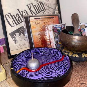 May include: A hand-painted ceramic ashtray with a black rim and a purple interior featuring a zebra stripe design. A silver sphere sits in the centre, with a red and orange line. A wooden stick is inserted into the sphere.