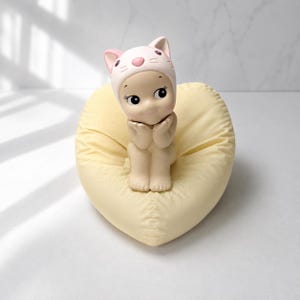 以下が含まれることがあります： ピンクの猫の帽子をかぶった小さな人形の置物。ハート型の黄色のクッションの上に座っています。人形は大きな目を持ち、手を顔の近くに置いています。クッションは柔らかく、質感のある生地で作られています。