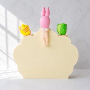 Puede incluir: Un letrero decorativo crema con forma de nube, adornado con un conejito rosa, un p&aacute;jaro amarillo y un caim&aacute;n verde. Estas figuras est&aacute;n colocadas en el borde del letrero, a&ntilde;adiendo un elemento l&uacute;dico y decorativo al dise&ntilde;o.