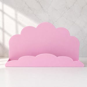 May include: A light pink wall-mounted shelf featuring a scalloped edge design that mimics the shape of a cloud. The shelf has a flat base and a tall, curved back panel with a smooth, matte finish, providing a simple storage surface.