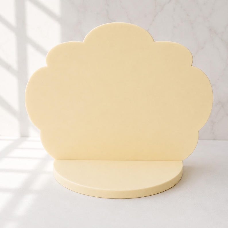 Puede incluir: Un expositor en forma de nube de color amarillo p&aacute;lido con una base semicircular. El expositor tiene un borde superior festoneado y est&aacute; hecho de un material s&oacute;lido y opaco. El fondo es una superficie blanca con un patr&oacute;n de m&aacute;rmol.