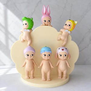 Puede incluir: Colecci&oacute;n de seis peque&ntilde;as figuras, cada una con un sombrero o gorro &uacute;nico. Las figuras desnudas, con caras pintadas, est&aacute;n dispuestas sobre una base en forma de nube de color amarillo claro. Los sombreros incluyen una rana, un conejo y un tigre.