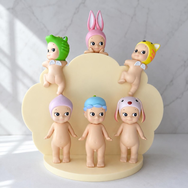 Puede incluir: Colecci&oacute;n de seis peque&ntilde;as figuras, cada una con un sombrero o gorro &uacute;nico. Las figuras desnudas, con caras pintadas, est&aacute;n dispuestas sobre una base en forma de nube de color amarillo claro. Los sombreros incluyen una rana, un conejo y un tigre.