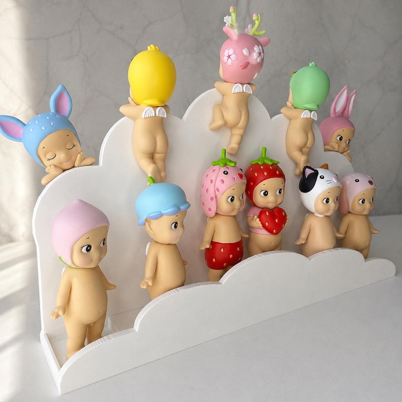 May include: A white cloud-shaped display shelf holding eleven small, nude baby figurines with various colorful headgear, including animal ears, fruit-themed hats, and floral designs. The figurines are arranged in two tiers on the shelf.