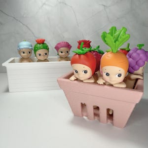 May include: A selection of small, quirky figurines, each with a distinctive vegetable or fruit-themed head covering. The figurines are in a white rectangular container and a pink berry basket. The heads include a cactus, rose, tomato, carrot, and grapes.
