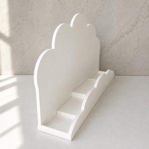 May include: A white display stand featuring a cloud-shaped back panel with a scalloped top edge. The base includes a tiered shelf structure with a matching scalloped front rail, providing multiple levels for organizing or showcasing small items.
