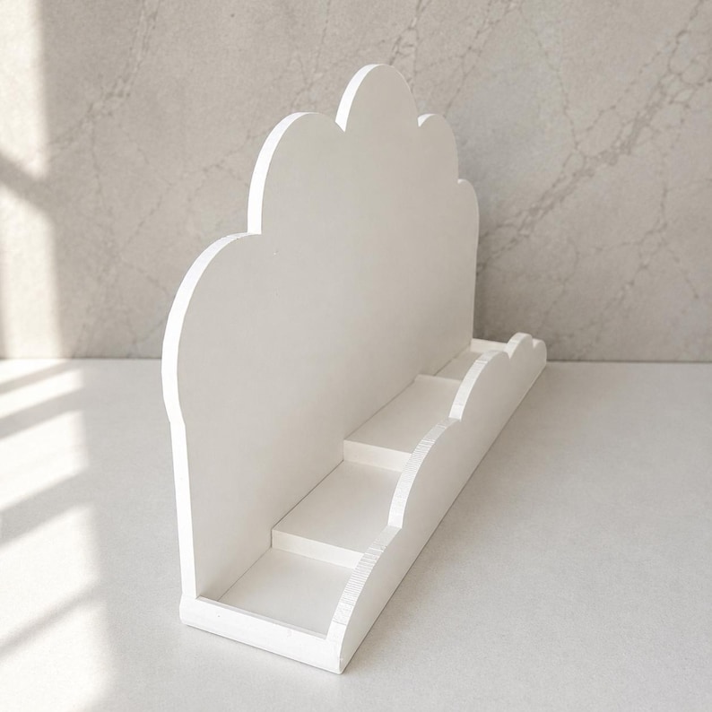 May include: A white display stand featuring a cloud-shaped back panel with a scalloped top edge. The base includes a tiered shelf structure with a matching scalloped front rail, providing multiple levels for organizing or showcasing small items.