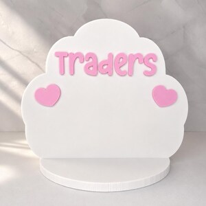 May include: A white cloud-shaped display stand featuring the pink text Traders centered at the top. Two pink heart-shaped accents are positioned on the left and right sides of the display. The stand includes a flat, semi-circular base for stability.