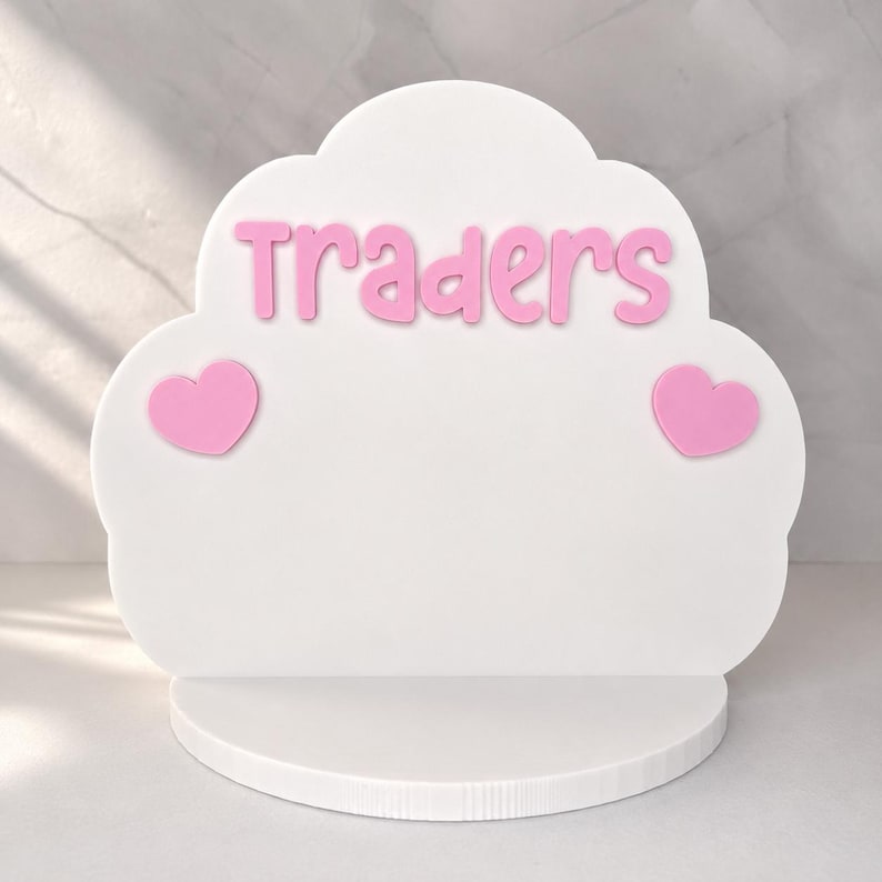 May include: A white cloud-shaped display stand featuring the pink text Traders centered at the top. Two pink heart-shaped accents are positioned on the left and right sides of the display. The stand includes a flat, semi-circular base for stability.
