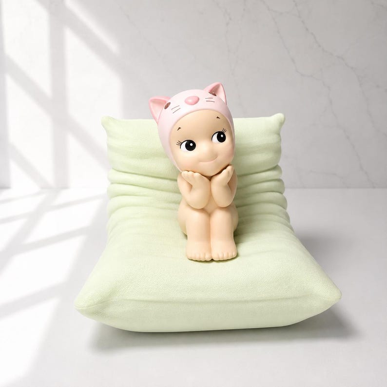 May include: A small, nude figurine wearing a pink cat-eared hat, seated on a light green pillow. The figurine has large, expressive eyes and is holding its hands near its face. The pillow has a soft, textured appearance.