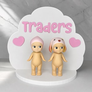 May include: A white cloud-shaped display stand featuring the pink text "Traders" and two pink hearts. Two small, nude, standing figurines are positioned on the base; one wears a pink pointed cap, and the other wears a pink and yellow dog-ear cap.