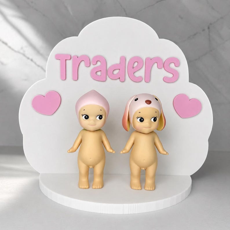 May include: A white cloud-shaped display stand featuring the pink text "Traders" and two pink hearts. Two small, nude, standing figurines are positioned on the base; one wears a pink pointed cap, and the other wears a pink and yellow dog-ear cap.