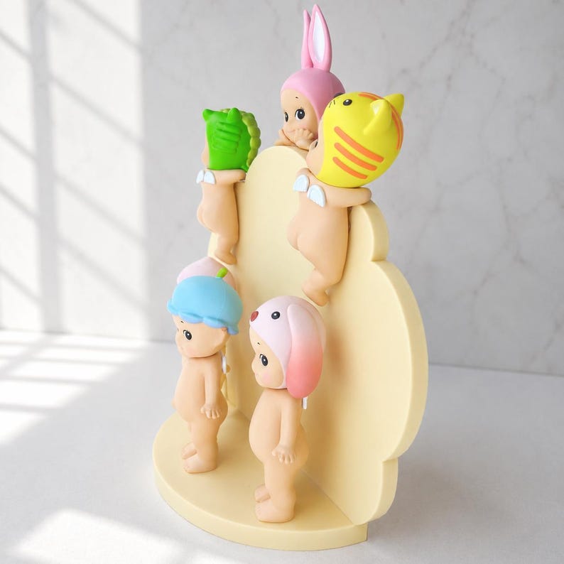 Puede incluir: Colecci&oacute;n de peque&ntilde;as figuras, cada una con un sombrero &uacute;nico con tem&aacute;tica de animales. Las figuras est&aacute;n dispuestas sobre una pantalla amarilla clara en forma de nube. Los sombreros incluyen un conejito rosa, un gato amarillo, un caim&aacute;n verde, un sombrero azul y un perro rosa.