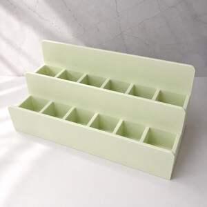 Puede incluir: Un organizador escalonado de color verde claro con dos niveles y seis compartimentos cuadrados en cada fila. La estructura est&aacute; hecha de un material s&oacute;lido y liso con acabado mate, dise&ntilde;ado con una disposici&oacute;n en niveles para organizar.