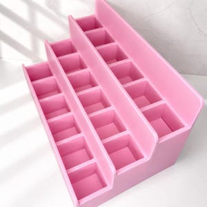 Puede incluir: Un organizador de pl&aacute;stico rosa de varios niveles con tres filas de compartimentos cuadrados. La estructura es escalonada, siendo la fila trasera la m&aacute;s alta y la delantera la m&aacute;s corta, lo que permite ver claramente cada secci&oacute;n de almacenamiento.