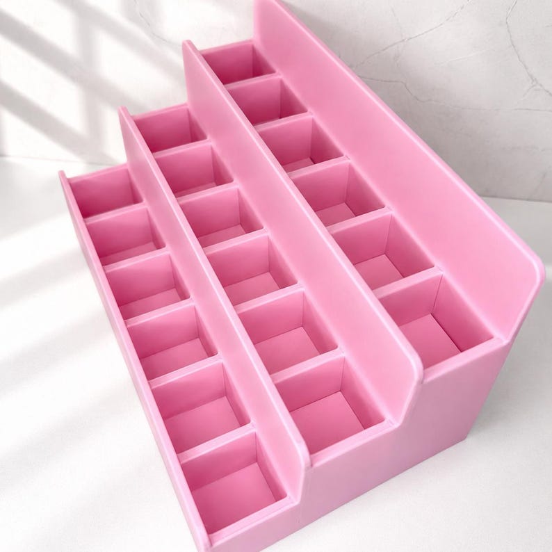 Puede incluir: Un organizador de pl&aacute;stico rosa de varios niveles con tres filas de compartimentos cuadrados. La estructura es escalonada, siendo la fila trasera la m&aacute;s alta y la delantera la m&aacute;s corta, lo que permite ver claramente cada secci&oacute;n de almacenamiento.