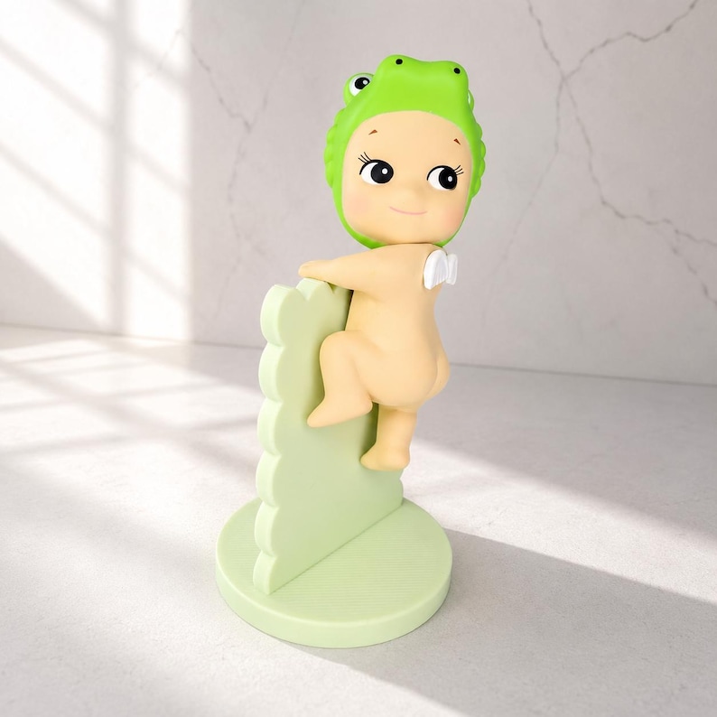 May include: A whimsical figurine of a nude baby wearing a green frog hat, climbing a light green scalloped wall. The figurine has a smiling face and small white wings. The base is a light green circle.