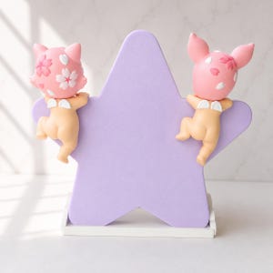 Op de afbeelding: Een decoratief stervormig beeldje in lavendel, ondersteund door een witte basis. Twee kleine figuren met roze hoofden, versierd met bloemmotieven en huidkleurige lichamen, zijn bevestigd aan de ster. De figuren hebben kleine witte vleugels.