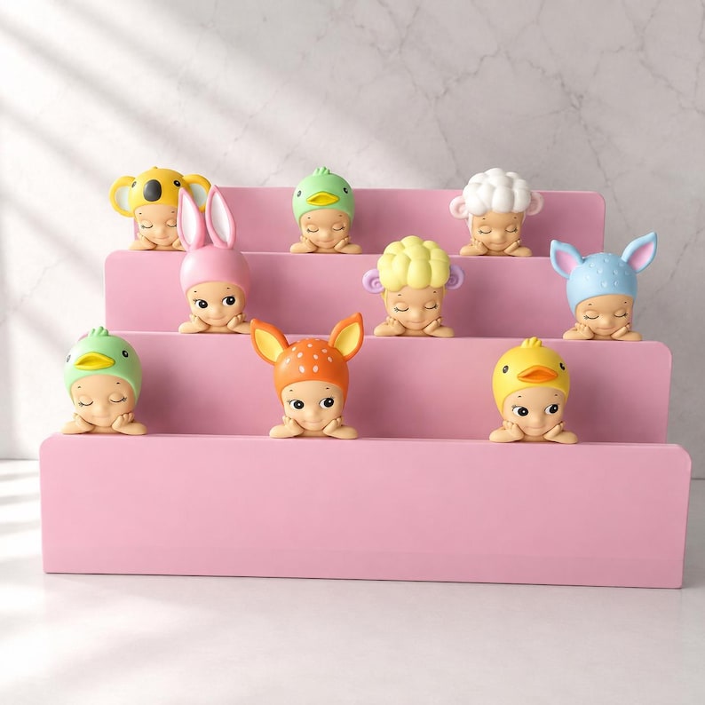 Puede incluir: Un expositor rosa de tres niveles con ocho peque&ntilde;as figuras. Cada figura presenta una cara de beb&eacute; con un gorro de tem&aacute;tica animal diferente, incluyendo un koala amarillo, un conejo rosa, un pato verde, una oveja blanca, un conejo azul, un p&aacute;jaro verde, un ciervo naranja y un pollito amarillo.