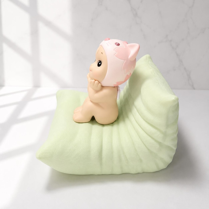 May include: A small, nude figurine wearing a pink cat-eared bonnet, seated on a light green, leaf-shaped pillow. The figurine has a serene expression, with its hands gently touching its face. The pillow has a soft, textured appearance.