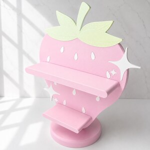 Puede incluir: Estanter&iacute;a rosa con forma de fresa de dos niveles. La estanter&iacute;a tiene un dise&ntilde;o de hojas verde claro en la parte superior y detalles blancos. La estanter&iacute;a est&aacute; dise&ntilde;ada para parecerse a una fresa, con detalles de semillas blancas.