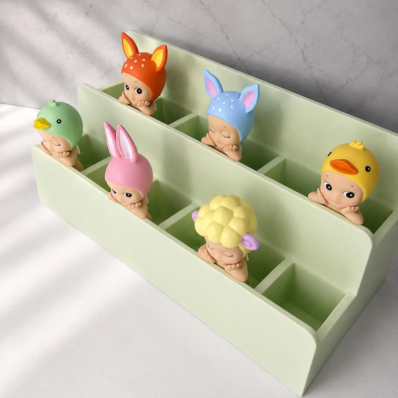 Puede incluir: Un organizador de almacenamiento de dos niveles en color verde claro con seis compartimentos, cada uno con una peque&ntilde;a figura que lleva un tocado de animal diferente, incluyendo un ciervo, un conejo azul, un p&aacute;jaro verde, un conejo rosa, una oveja amarilla y un pato.
