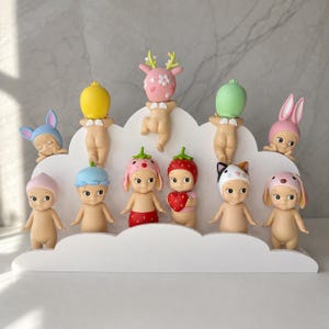 May include: A collection of twelve small, nude baby figurines with various animal and fruit-themed headgear, including a deer, strawberry, and cat, displayed on a white, cloud-shaped tiered stand. The figures feature small wings on their backs.