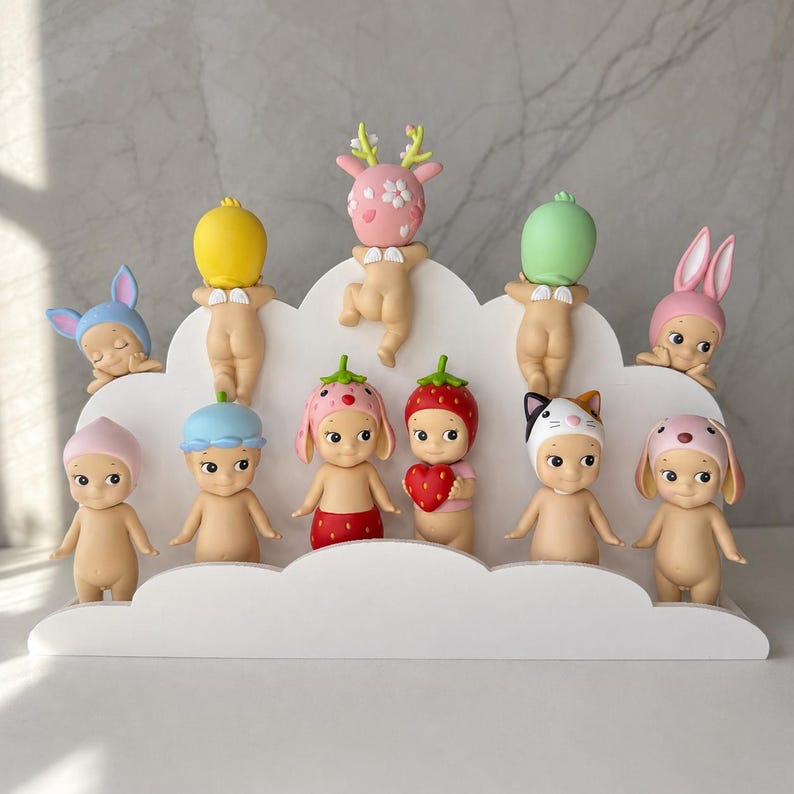 May include: A collection of twelve small, nude baby figurines with various animal and fruit-themed headgear, including a deer, strawberry, and cat, displayed on a white, cloud-shaped tiered stand. The figures feature small wings on their backs.