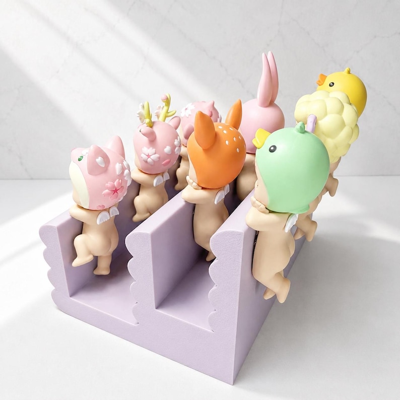 May include: A collection of whimsical figurines, each with a unique animal-themed head, including a bunny, deer, and duck. They are arranged in a lavender-colored, stepped display unit. The figurines have small wings and are in various pastel colors.