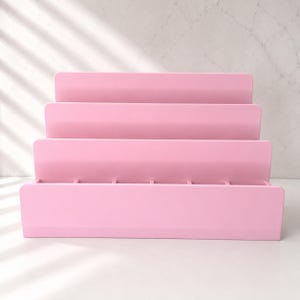 Puede incluir: Un expositor escalonado de color rosa claro con cuatro estantes horizontales dispuestos en forma de escalera. La estructura est&aacute; construida con paneles s&oacute;lidos e incluye peque&ntilde;os divisores verticales en el estante base para crear ranuras.
