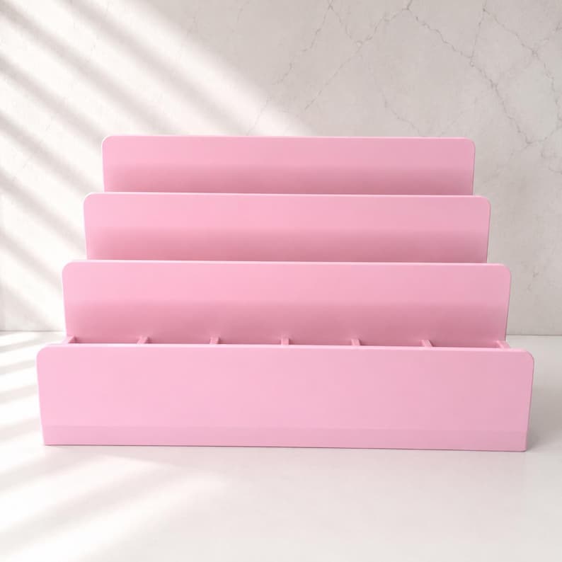 Puede incluir: Un expositor escalonado de color rosa claro con cuatro estantes horizontales dispuestos en forma de escalera. La estructura est&aacute; construida con paneles s&oacute;lidos e incluye peque&ntilde;os divisores verticales en el estante base para crear ranuras.