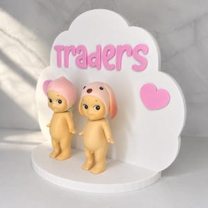 May include: A white cloud-shaped display stand featuring the pink text "traders" and a pink heart. Two small, light-skinned figurines stand on the base; one wears a pink peach-shaped hat, and the other wears a pink dog-eared hat.