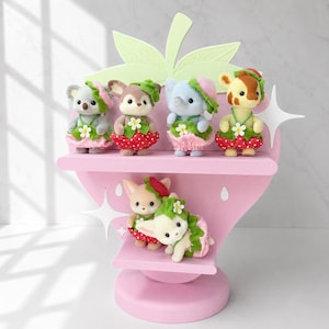 Puede incluir: Una estanter&iacute;a rosa con forma de fresa exhibe seis figuras de animales en miniatura. Las figuras incluyen un koala, un ciervo, un elefante y una jirafa, cada uno con un atuendo con tem&aacute;tica de fresa. La estanter&iacute;a tiene hojas verdes y acentos de estrellas blancas.