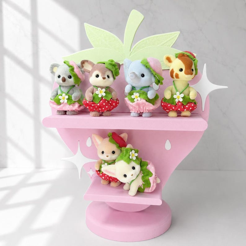 Puede incluir: Una estanter&iacute;a rosa con forma de fresa exhibe seis figuras de animales en miniatura. Las figuras incluyen un koala, un ciervo, un elefante y una jirafa, cada uno con un atuendo con tem&aacute;tica de fresa. La estanter&iacute;a tiene hojas verdes y acentos de estrellas blancas.