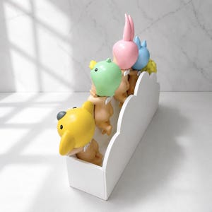 Peut inclure: Une pi&egrave;ce d&eacute;corative fantaisiste avec un contenant blanc en forme de nuage contenant des figurines avec des t&ecirc;tes en forme de fruits jaunes, vertes, roses et bleues. Les figurines semblent grimper sur la structure nuageuse.
