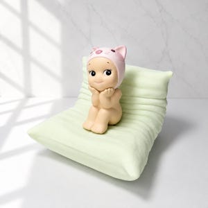 May include: A small, beige figurine of a seated figure wearing a pink cat-eared hat, resting on a light green pillow. The figurine has large, black eyes and is posed with hands under its chin. The pillow has a ribbed texture.