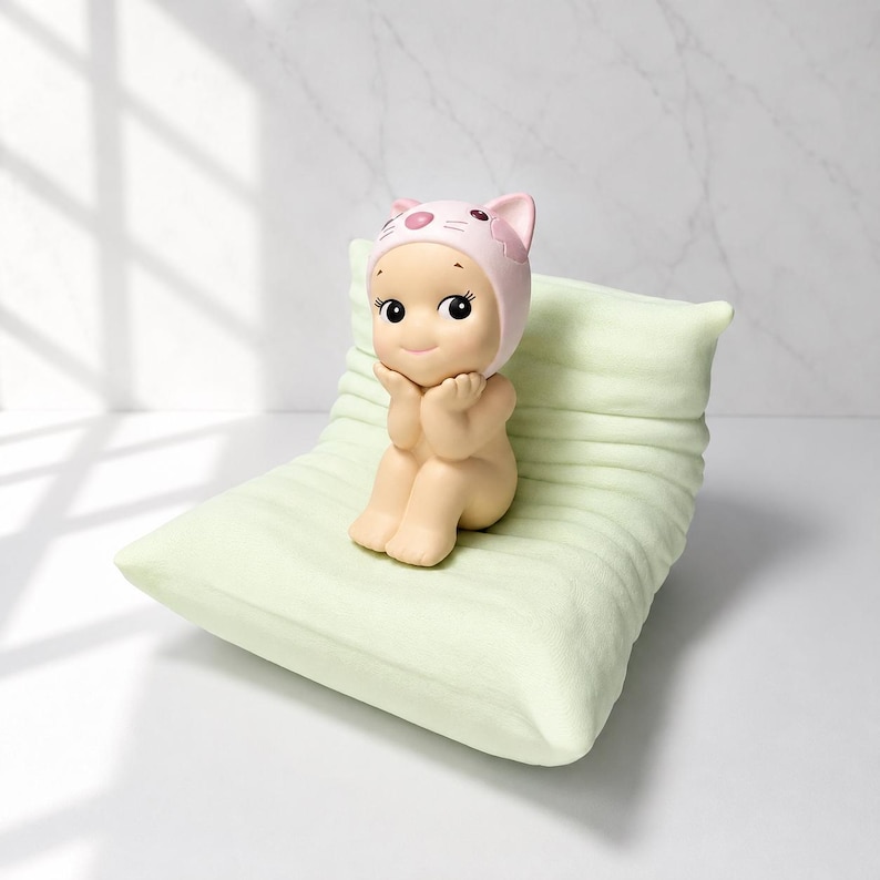May include: A small, beige figurine of a seated figure wearing a pink cat-eared hat, resting on a light green pillow. The figurine has large, black eyes and is posed with hands under its chin. The pillow has a ribbed texture.