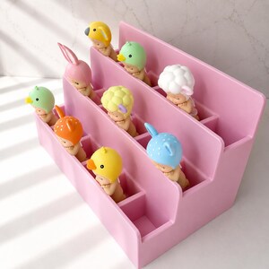 Puede incluir: Un expositor de pl&aacute;stico rosa de varios niveles con m&uacute;ltiples compartimentos, cada uno con una peque&ntilde;a figura de beb&eacute; que lleva un gorro con forma de animal, incluyendo un pato amarillo, un conejo rosa, un p&aacute;jaro verde, una oveja blanca, un ciervo naranja y un oso azul.