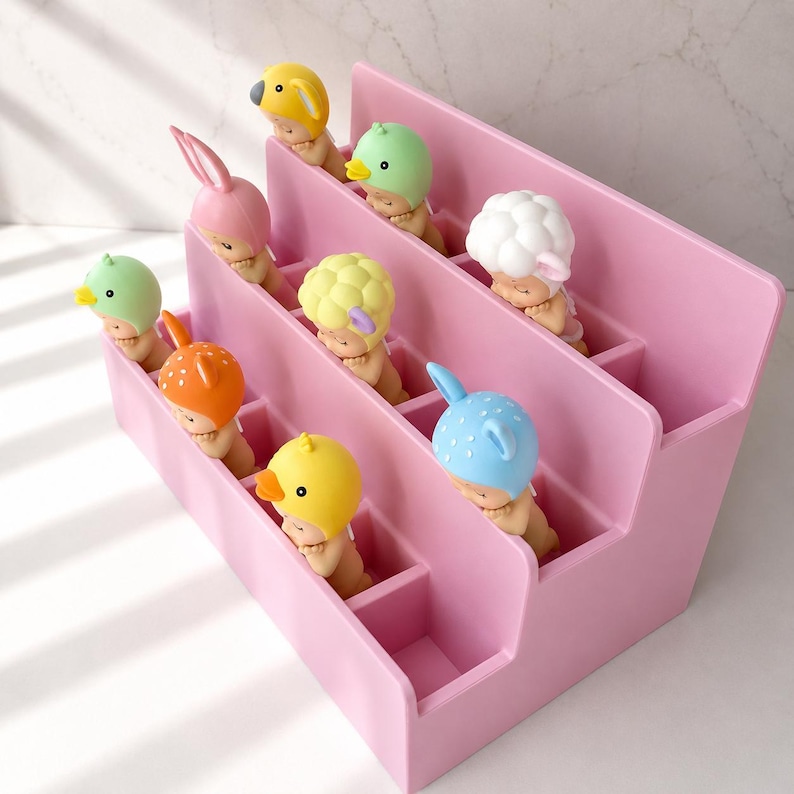 Puede incluir: Un expositor de pl&aacute;stico rosa de varios niveles con m&uacute;ltiples compartimentos, cada uno con una peque&ntilde;a figura de beb&eacute; que lleva un gorro con forma de animal, incluyendo un pato amarillo, un conejo rosa, un p&aacute;jaro verde, una oveja blanca, un ciervo naranja y un oso azul.