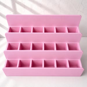 Puede incluir: Un expositor escalonado de color rosa claro con tres filas horizontales, cada una dividida en seis compartimentos cuadrados individuales. La estructura es abierta, cuenta con un panel trasero s&oacute;lido y un acabado mate liso para organizar objetos.