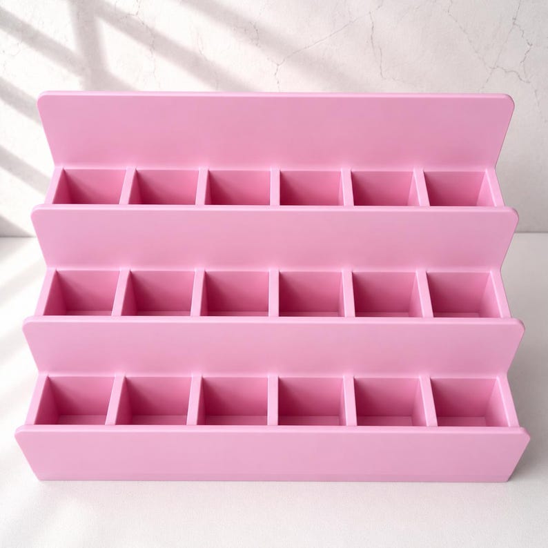 Puede incluir: Un expositor escalonado de color rosa claro con tres filas horizontales, cada una dividida en seis compartimentos cuadrados individuales. La estructura es abierta, cuenta con un panel trasero s&oacute;lido y un acabado mate liso para organizar objetos.