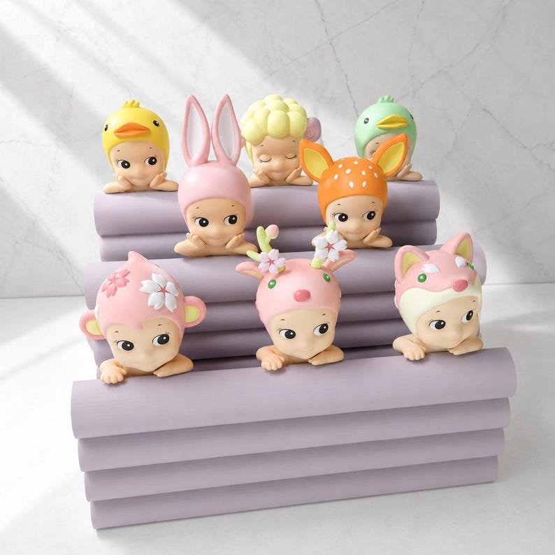 May include: A collection of small figurines, each with a unique animal-themed hat. The figurines are in various colors, including yellow, pink, orange, and green. The figurines are arranged on a stack of lavender-colored blocks.